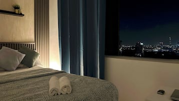 Een hotelkamer met een bed, nachtkastje, raam met uitzicht over de stad en een klok aan de muur.
