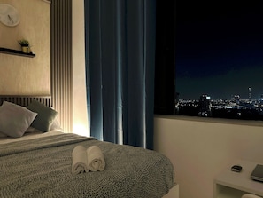 A hotel room with a bed, a bedside table, a window with a city view, and a clock on the wall. - Pathfinder Partition Room in Tecom (Dubai)