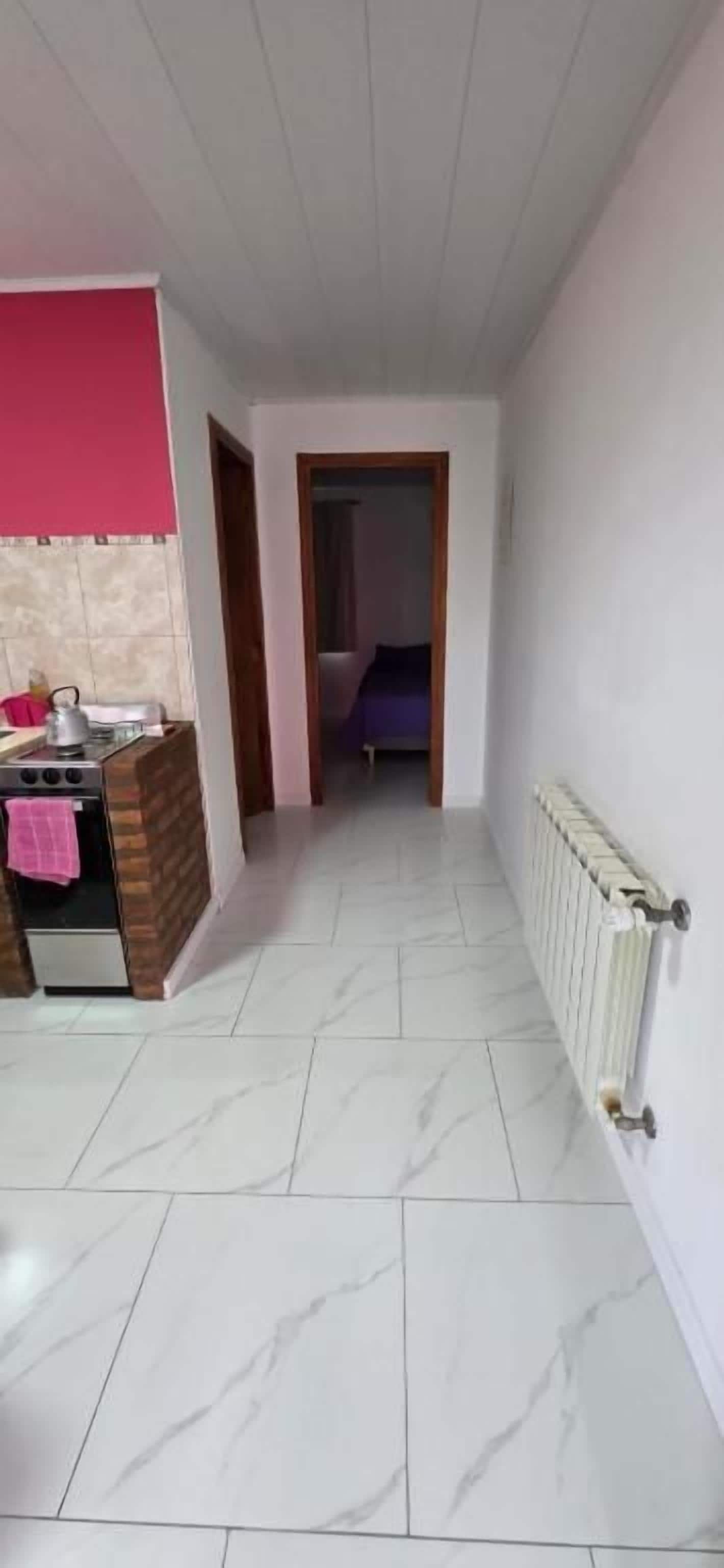 Corredor estreito com área de cozinha à esquerda, radiador à direita e uma cama visível por uma porta aberta no final.