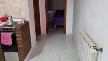 Corredor estreito com área de cozinha à esquerda, radiador à direita e uma cama visível por uma porta aberta no final.