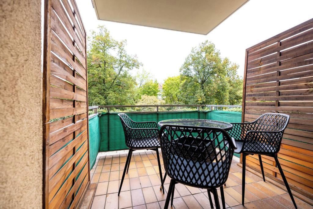 Ein Balkon mit Tisch und Stühlen, ein grüner Zaun und Bäume im Hintergrund.