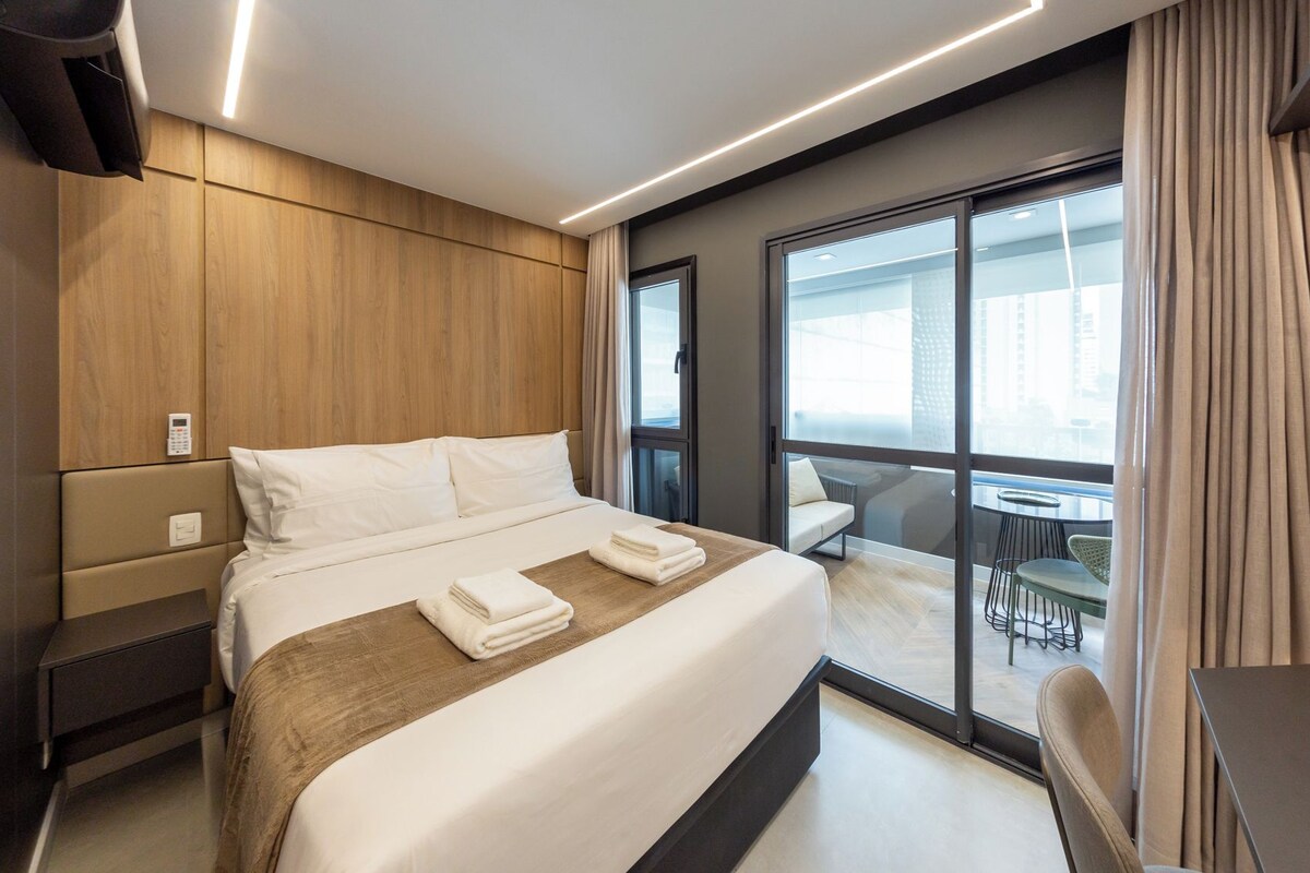 A modern hotel room with a large bed, a bedside table, a control panel, and a view of the balcony area.