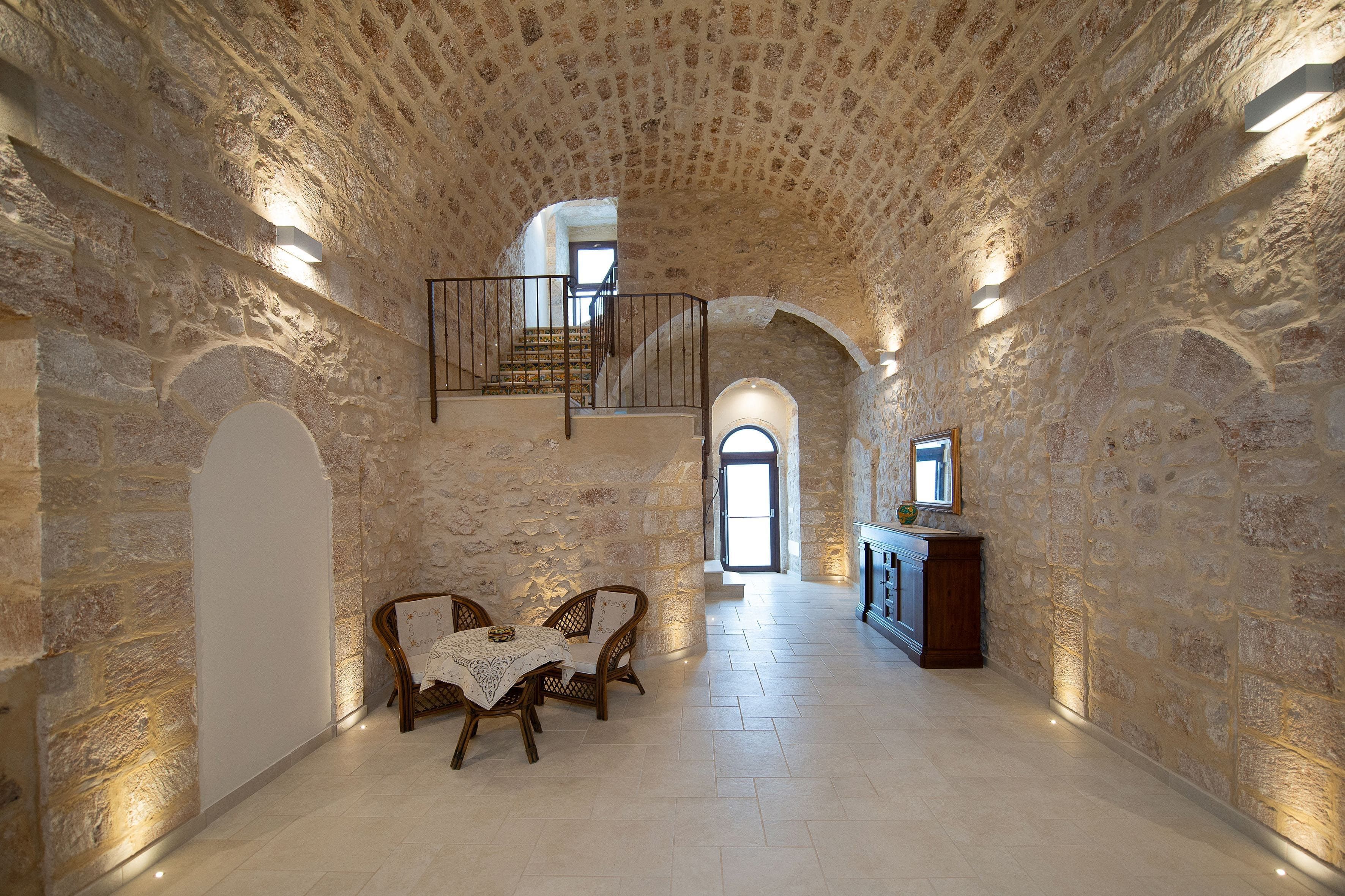 A stone-walled hallway with a staircase leading to an upper level, a table with chairs, and a cabinet.