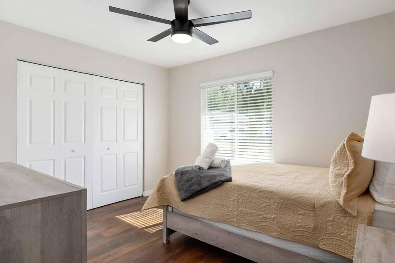 Une chambre à coucher avec un lit, un ventilateur de plafond, une fenêtre avec des stores et une porte.
