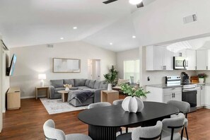 A modern living room with a grey sectional sofa, a round black table with chairs, a flat-screen TV mounted on the wall, and a kitchen area with white cabinets and appliances.