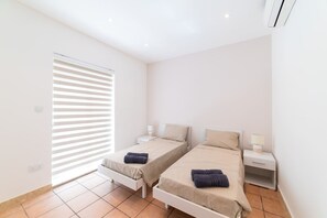 Quarto com duas camas, janela com persianas e mesa de cabeceira com abajur.