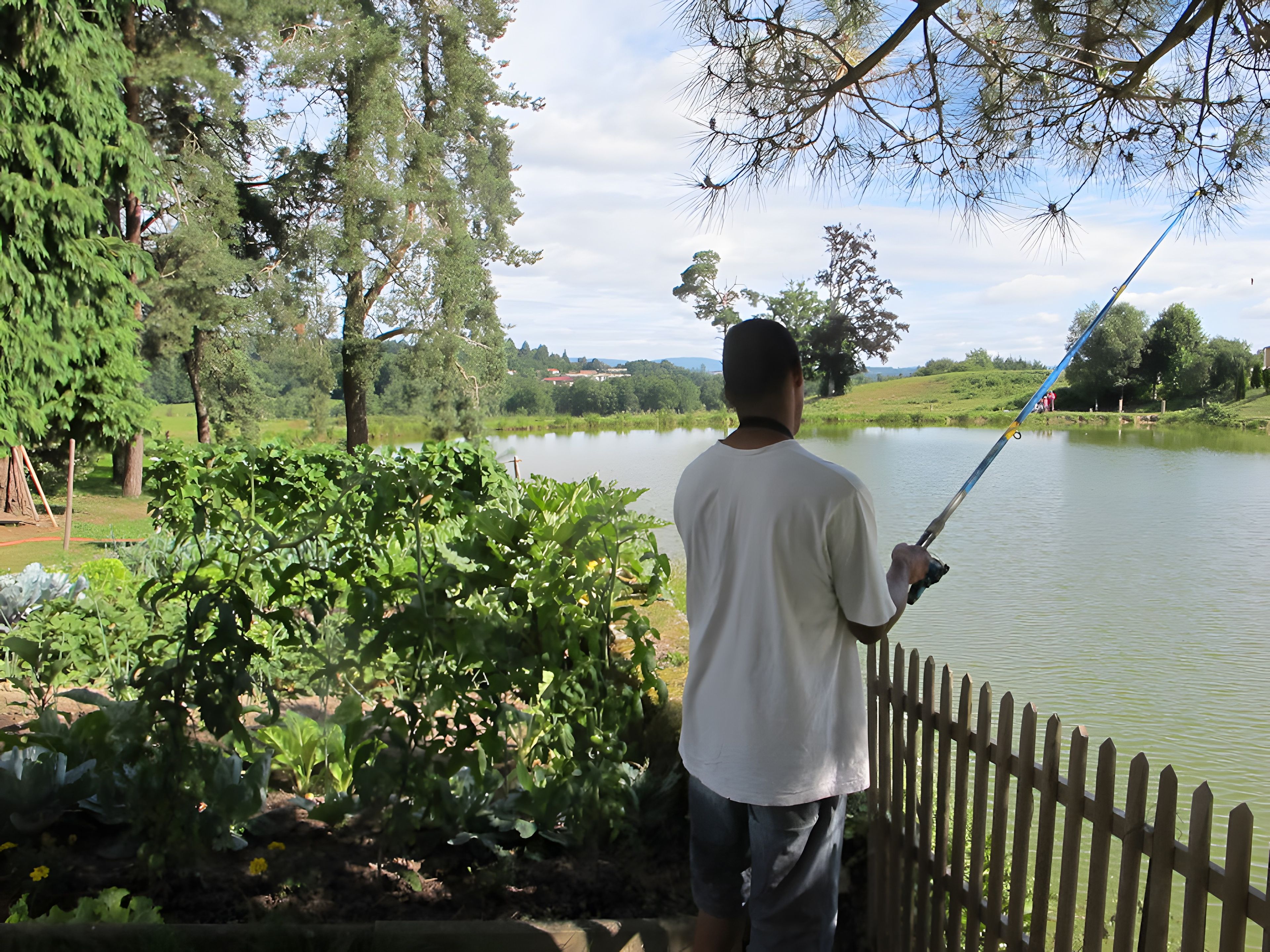 Une personne est en train de pêcher au bord d’un lac paisible, avec une clôture en bois au premier plan.