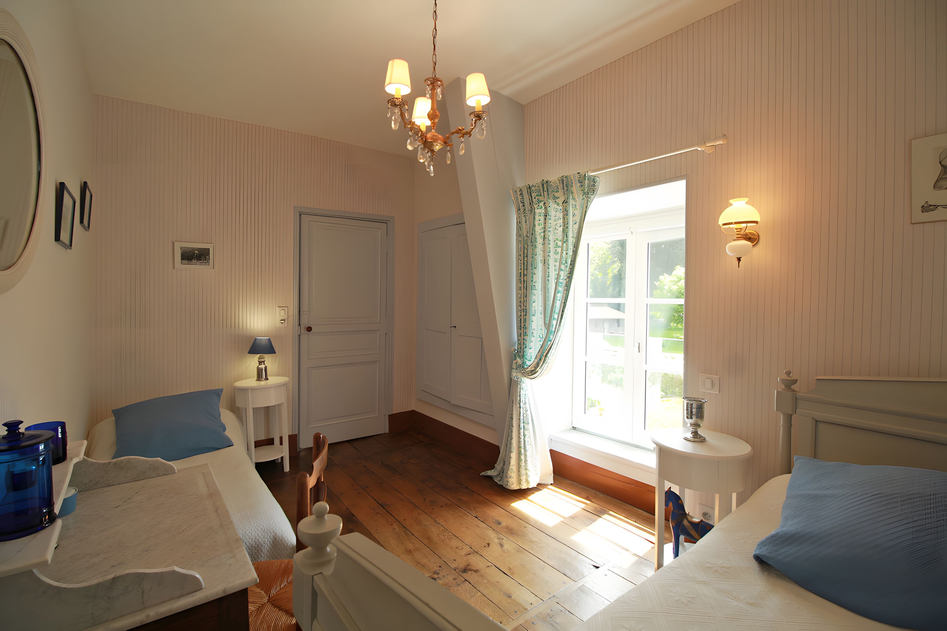 Et hotelværelse med trægulv, seng, skrivebord med lampe, stol, skab og et vindue med gardiner.