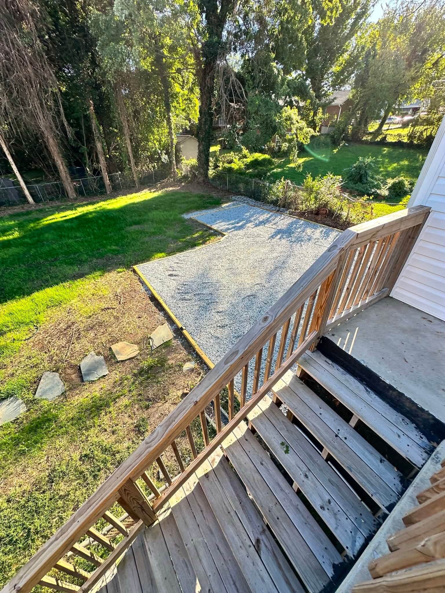 Une terrasse en bois avec une rambarde en métal, qui mène à un espace recouvert de gravier, agrémenté de gazon et d’arbres.