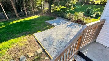 Une terrasse en bois avec une rambarde en métal, qui mène à un espace recouvert de gravier, agrémenté de gazon et d’arbres.