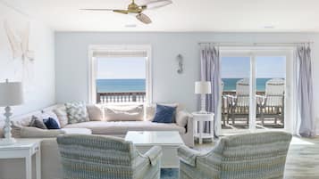 A living room with a sofa, two armchairs, a coffee table, a ceiling fan, and a view of the ocean through large windows.