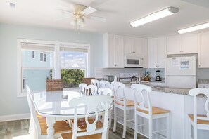 Une cuisine avec des meubles blancs, une table à manger avec des chaises, un ventilateur de plafond et, par les fenêtres, une vue sur une maison bleue.