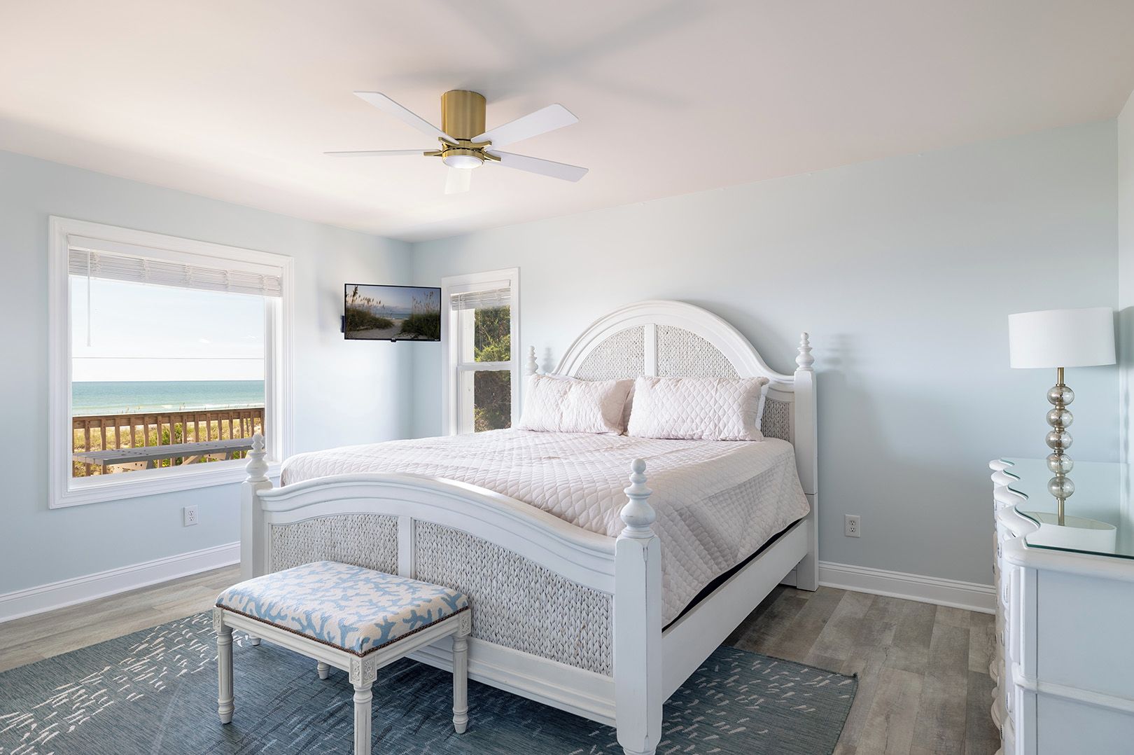A bedroom with a bed, a bench, a ceiling fan, a TV, and a view of the beach.
