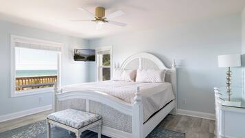A bedroom with a bed, a bench, a ceiling fan, a TV, and a view of the beach.