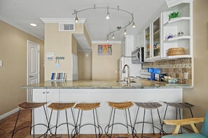 A kitchen with a central island, bar stools, a microwave, and a coffee machine. - Seaside Treasure-Top Floor Condo Pool & Beach View (Galveston)