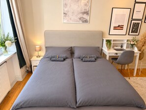 Una cama doble con ropa de cama gris, una mesita de noche con lámpara, un escritorio con silla y cuadros en la pared.