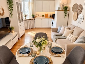 Una cocina moderna con armarios blancos, un comedor con una mesa redonda para cuatro personas, y una sala de estar acogedora con un sofá y cojines.