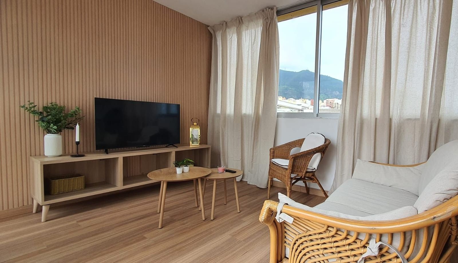 Ein Zimmer mit Fernseher auf einem Holzboden, einem Korbssessel, einem kleinen Tisch und Blick ins Freie durch ein Fenster.