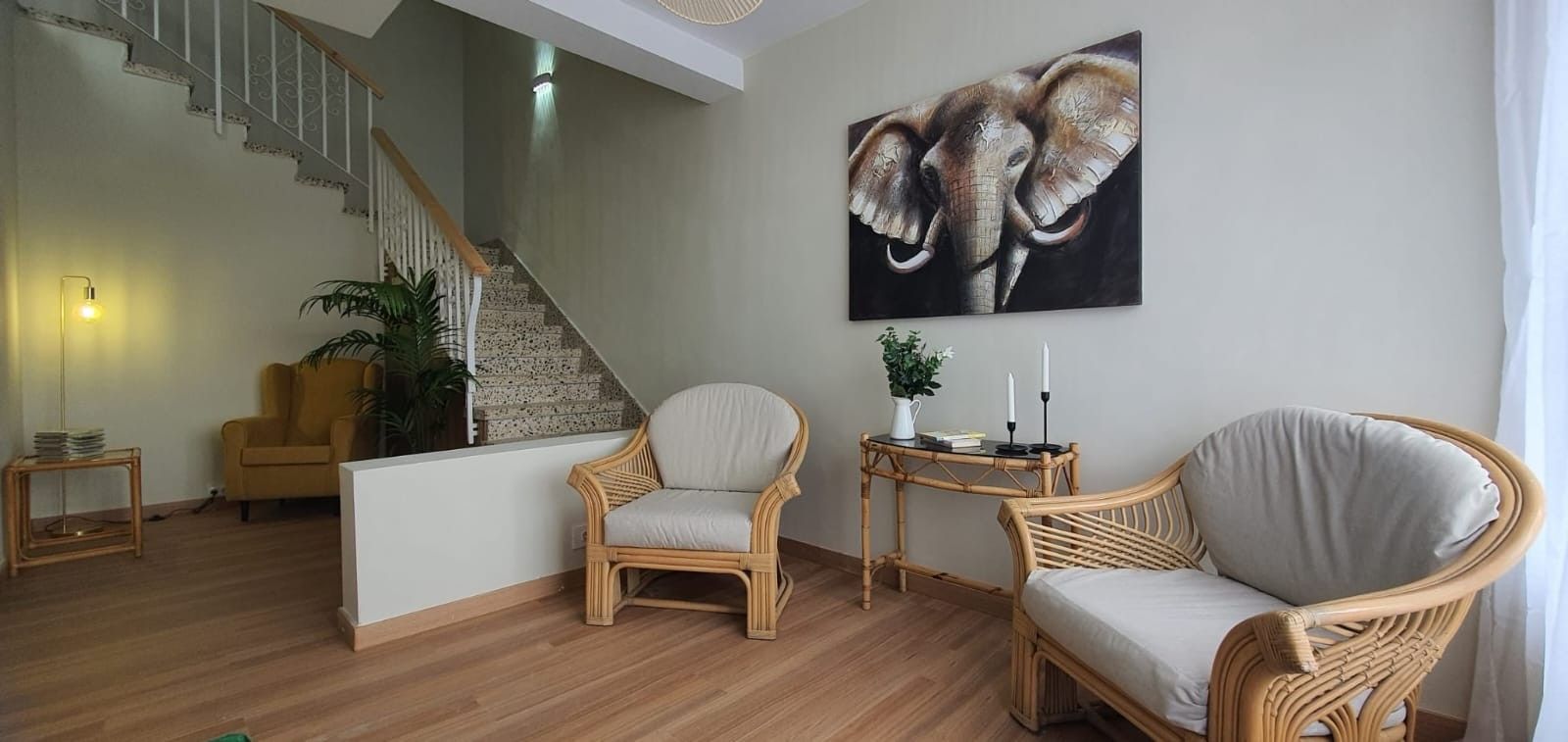 Ein Wohnzimmer mit Treppe, zwei Korbstühle, einem kleinen Tisch mit Kerzen und einem großen Elefantenbild an der Wand.