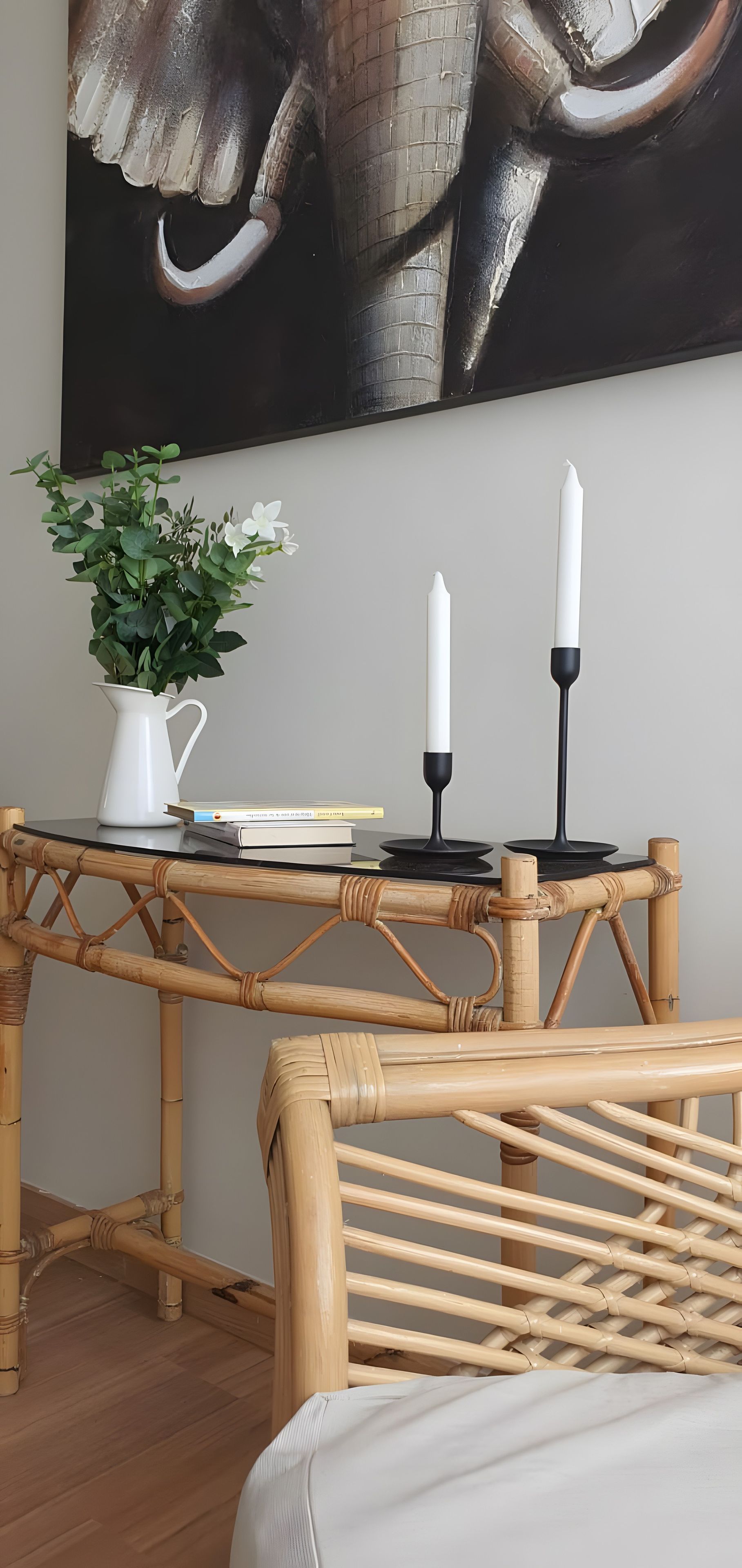 A room with a woven bamboo table, a white vase with green foliage, a book, and two white candles on black holders.