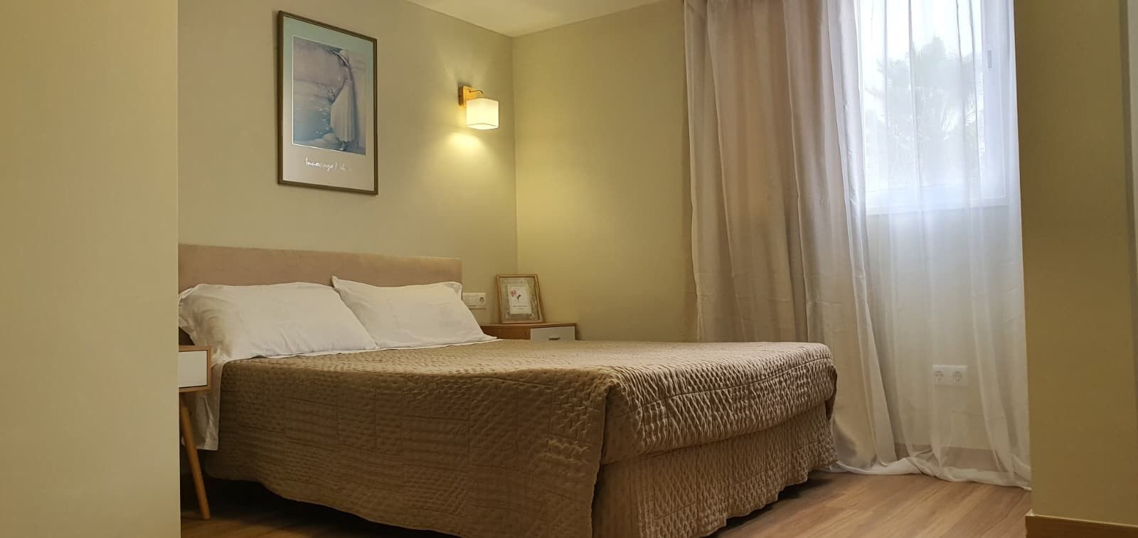 Habitación de hotel con cama, mesita de noche, un cuadro en la pared y ventana con cortinas traslúcidas.