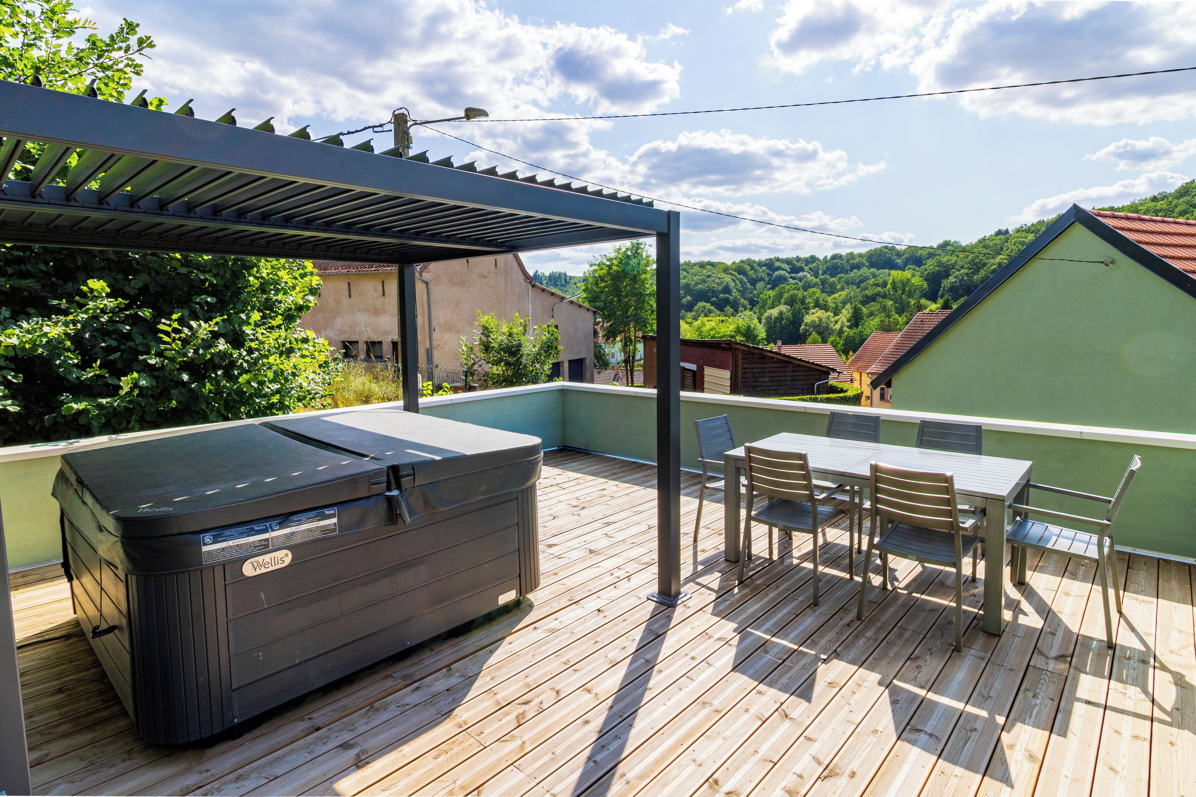 Une terrasse en bois avec un jacuzzi, du mobilier d’extérieur et une vue sur un bâtiment verdoyant et des arbres.