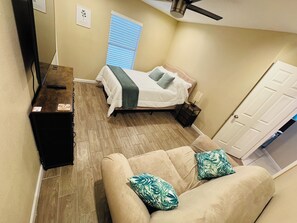 A bedroom with a bed, a bedside table, a nightstand, a TV, a ceiling fan, a window with blinds, and a door. - 30+ Night 4BR Near Lackland & SeaWorld (San Antonio)