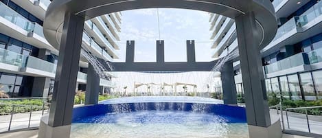 Un bâtiment moderne doté d’un élément aquatique central, de nombreux balcons et surplombant un ciel dégagé.