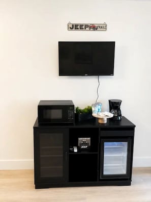 Un mobile porta TV nero con televisore montato sopra, un forno a microonde, una macchina del caffè e un piccolo frigorifero.