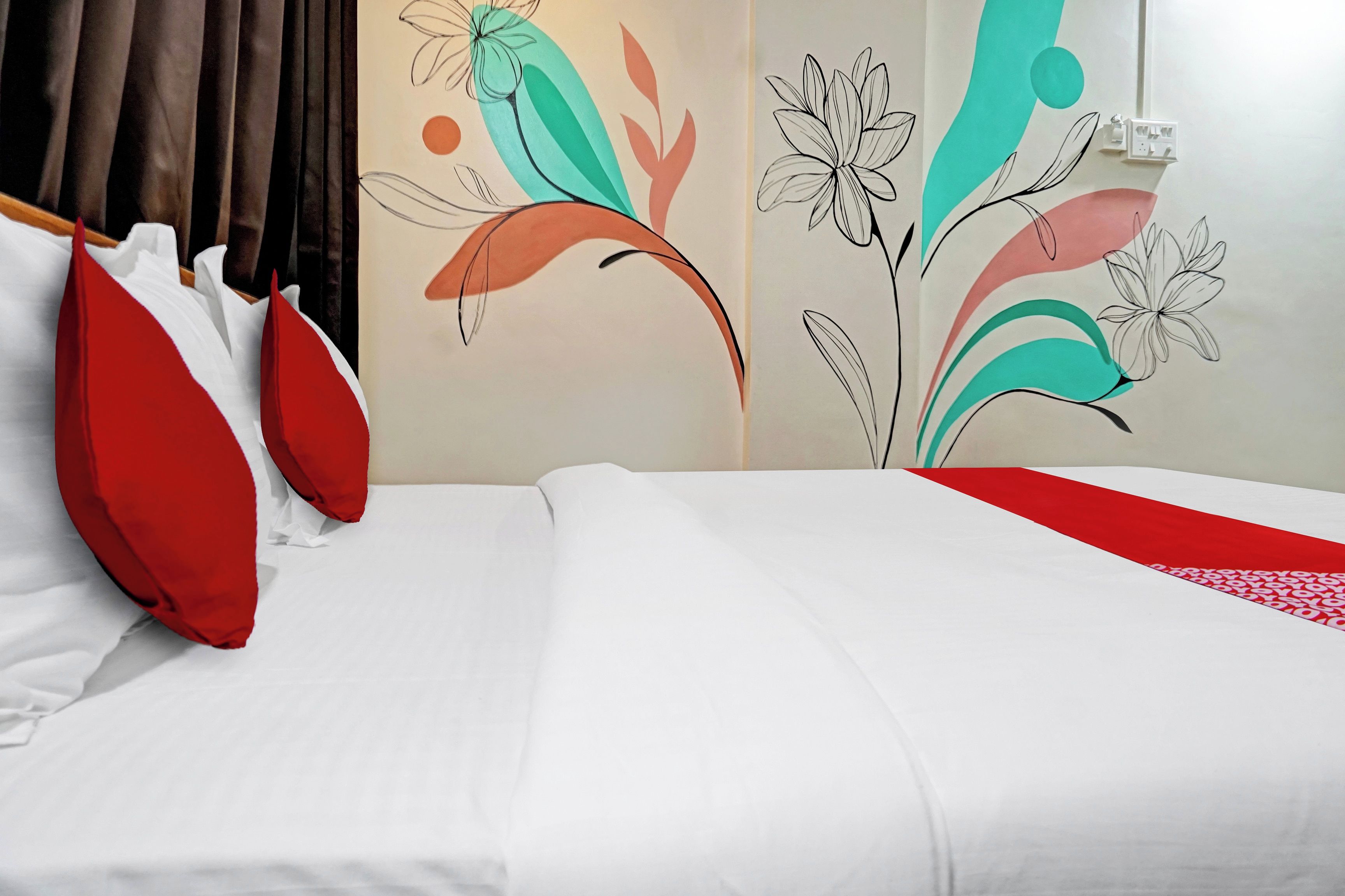 A hotel room with a bed, red and white pillows, and a wall with abstract floral wall art.