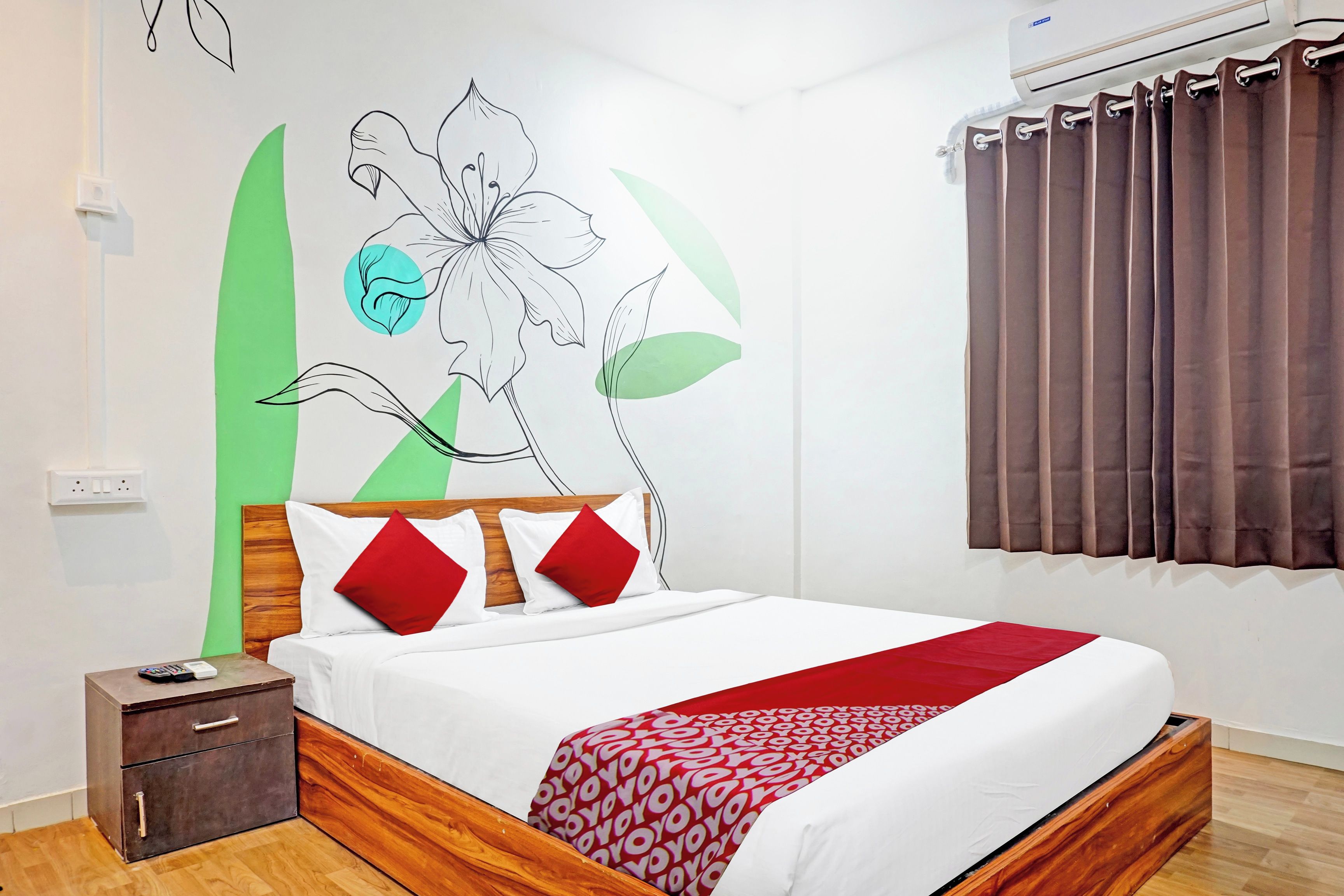 Habitación de hotel con cama, mesita de noche, cómoda de madera, pared con un mural floral y ventana con cortinas.