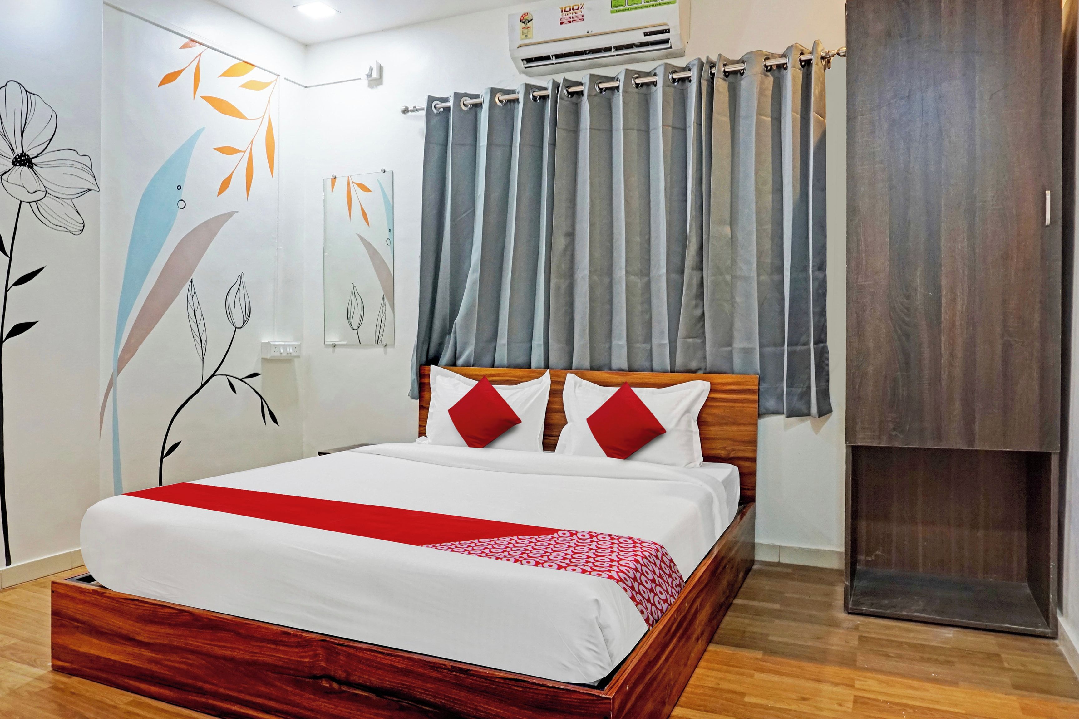 A hotel room with a wooden bed, a red and white bedspread, grey curtains, and a wall with floral artwork.