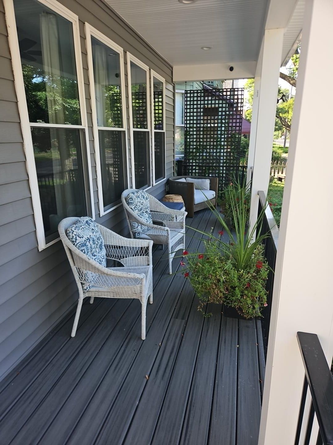 Een overdekt terras met twee rieten stoelen en een kleine bank, een plant in pot en een zwart metalen hek.