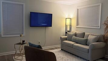 Un salon avec un téléviseur à écran plat fixé au mur, un canapé, une petite table d’appoint avec une lampe et un fauteuil.