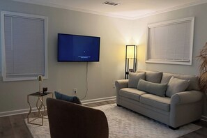 Un salon avec un téléviseur à écran plat fixé au mur, un canapé, une petite table d’appoint avec une lampe et un fauteuil.