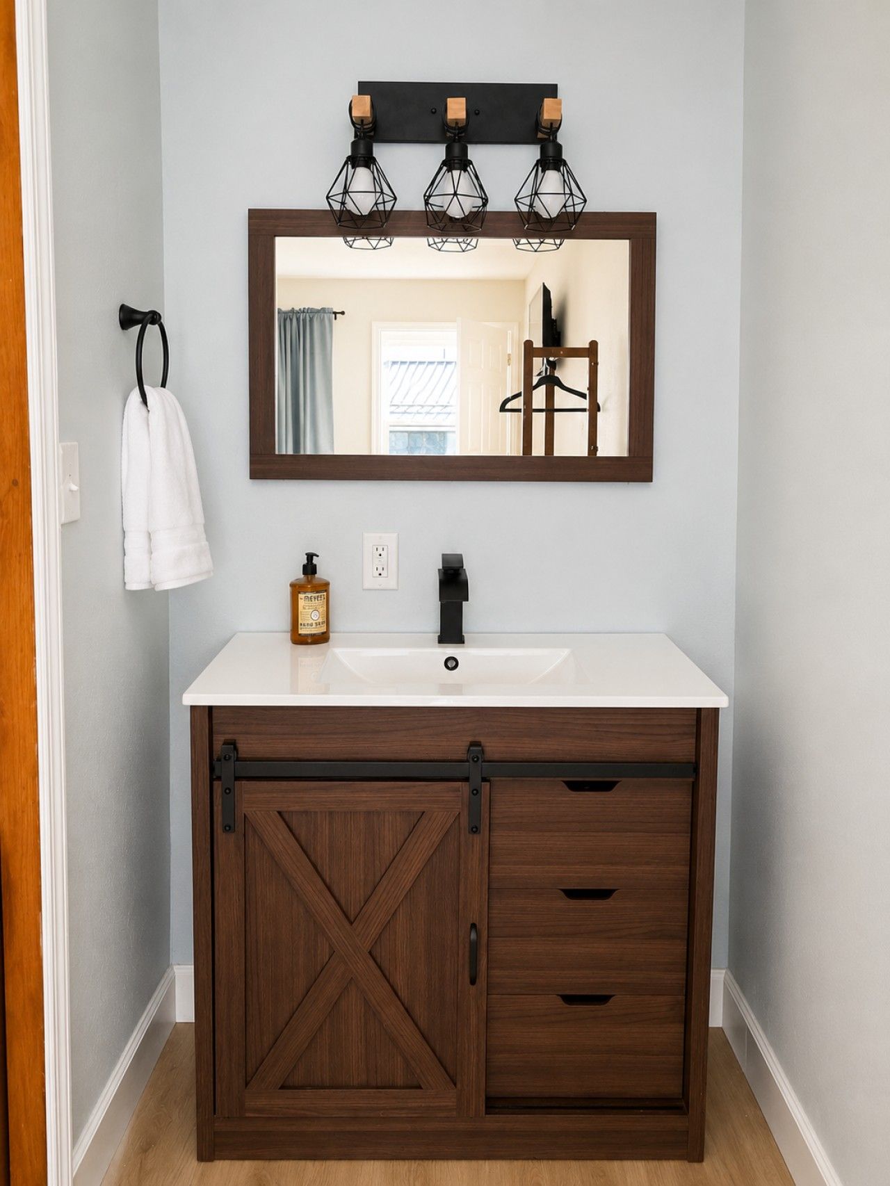 Un baño con un lavamanos blanco, un mueble de madera con diseño de cruz, un perchero y un espejo con marco negro.