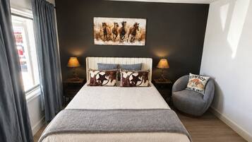 A bedroom with a bed, two bedside tables, a chair, a wall-mounted light fixture, and a painting.