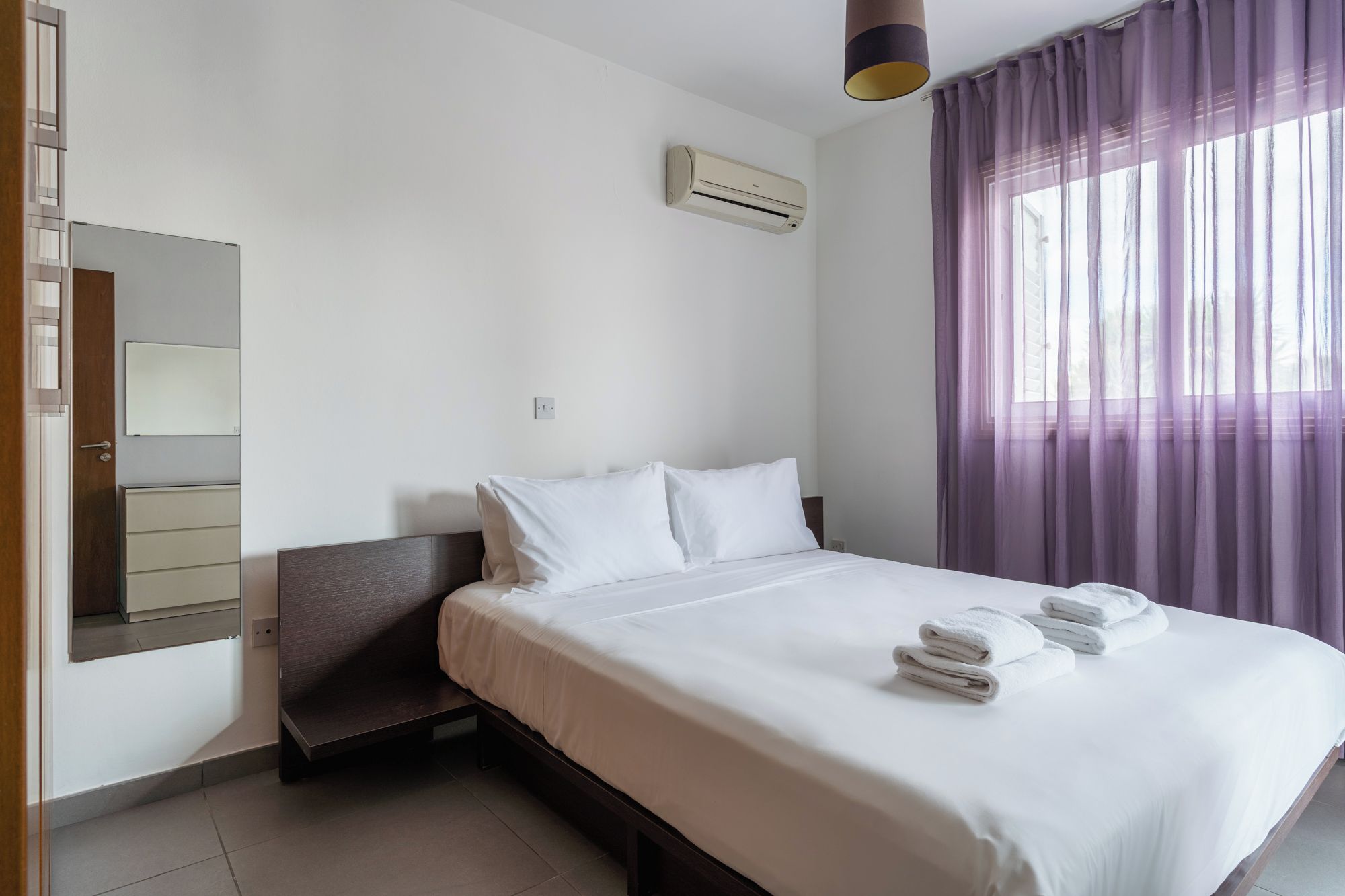 A hotel room with a bed, a bedside table, a mirror, an air conditioning unit, and a window with curtains.