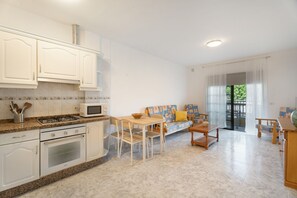 Een moderne keuken met witte kasten, een magnetron en een gasfornuis. Er is een eethoek met een houten tafel en stoelen, plus een bank met kussens. De kamer heeft een groot raam met gordijnen.