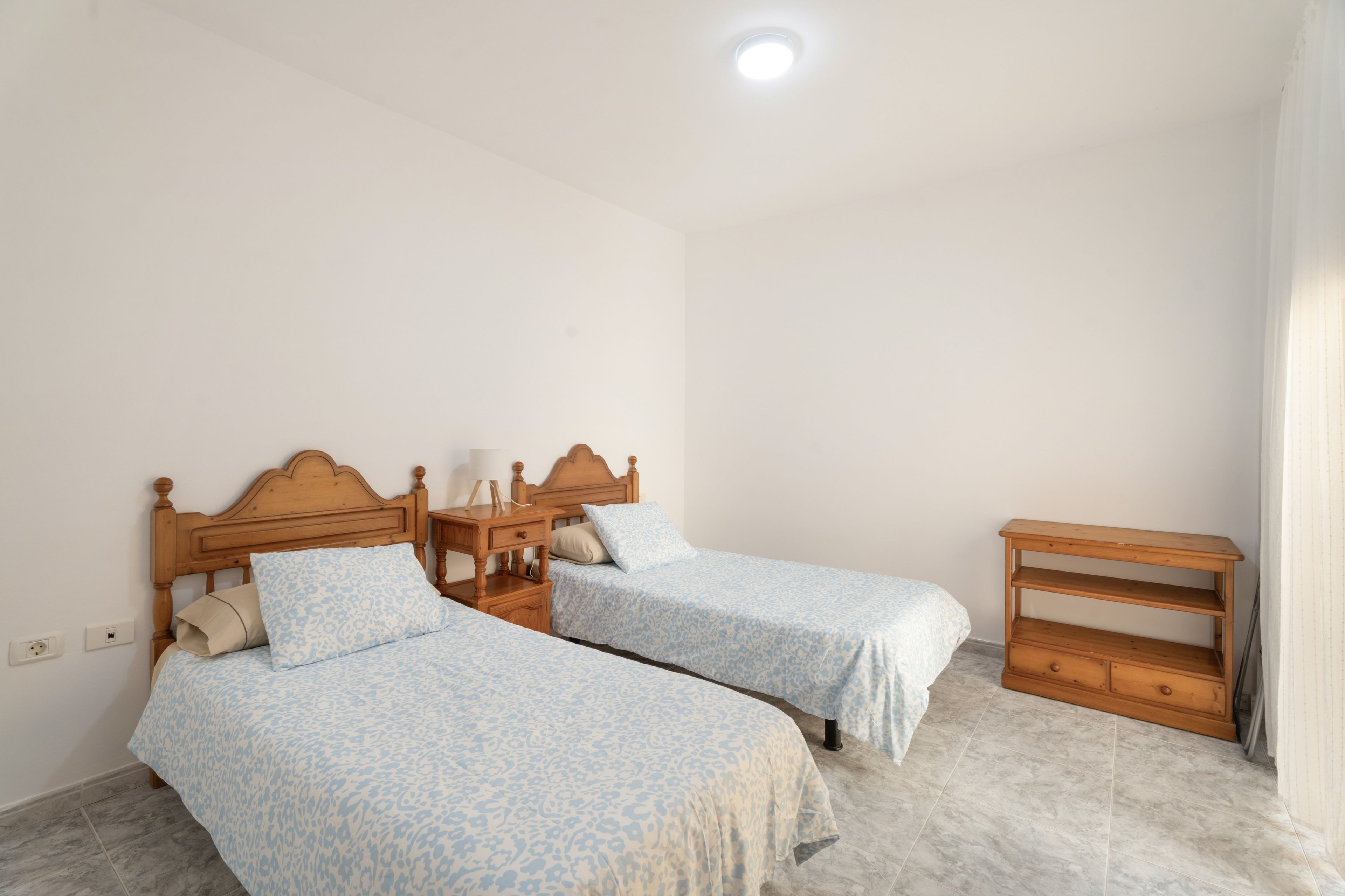 Un dormitorio con dos camas de madera, una mesita de noche, un aparador de madera y una lámpara en el techo.