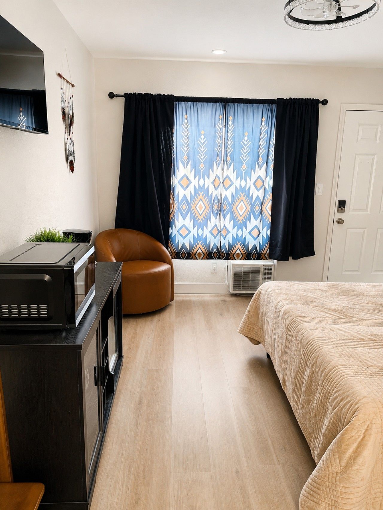 A compact hotel room with a bed, a brown leather chair, a desk with a computer, a window with patterned curtains, and a ceiling light fixture.