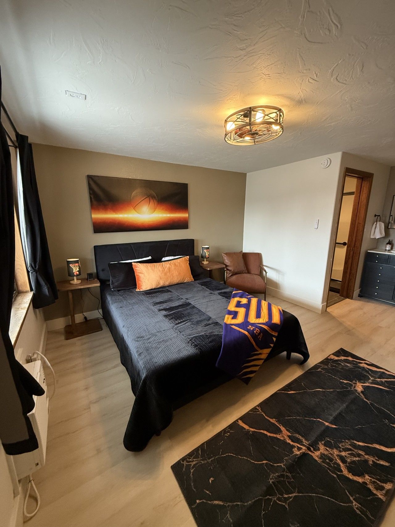 A bedroom with a bed, a nightstand, a lamp, a chair, a painting, and a bathroom visible through an open door.