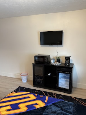 A room with a television mounted on the wall, a black entertainment unit with a microwave, a coffee maker, and a small refrigerator. There is a basketball jersey on the floor.