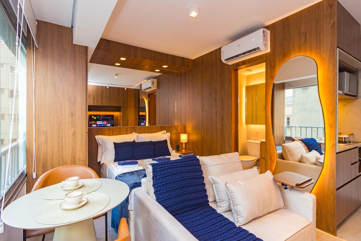 A modern hotel room with a bed, a seating area with a table and chairs, a large mirror, and an air conditioning unit.