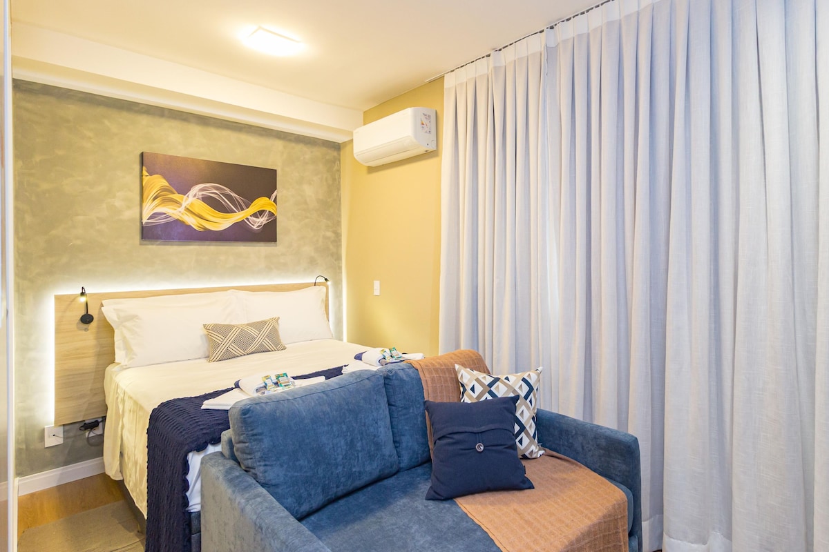 A hotel room with a bed, a sofa, a wall-mounted air conditioner, and a painting.