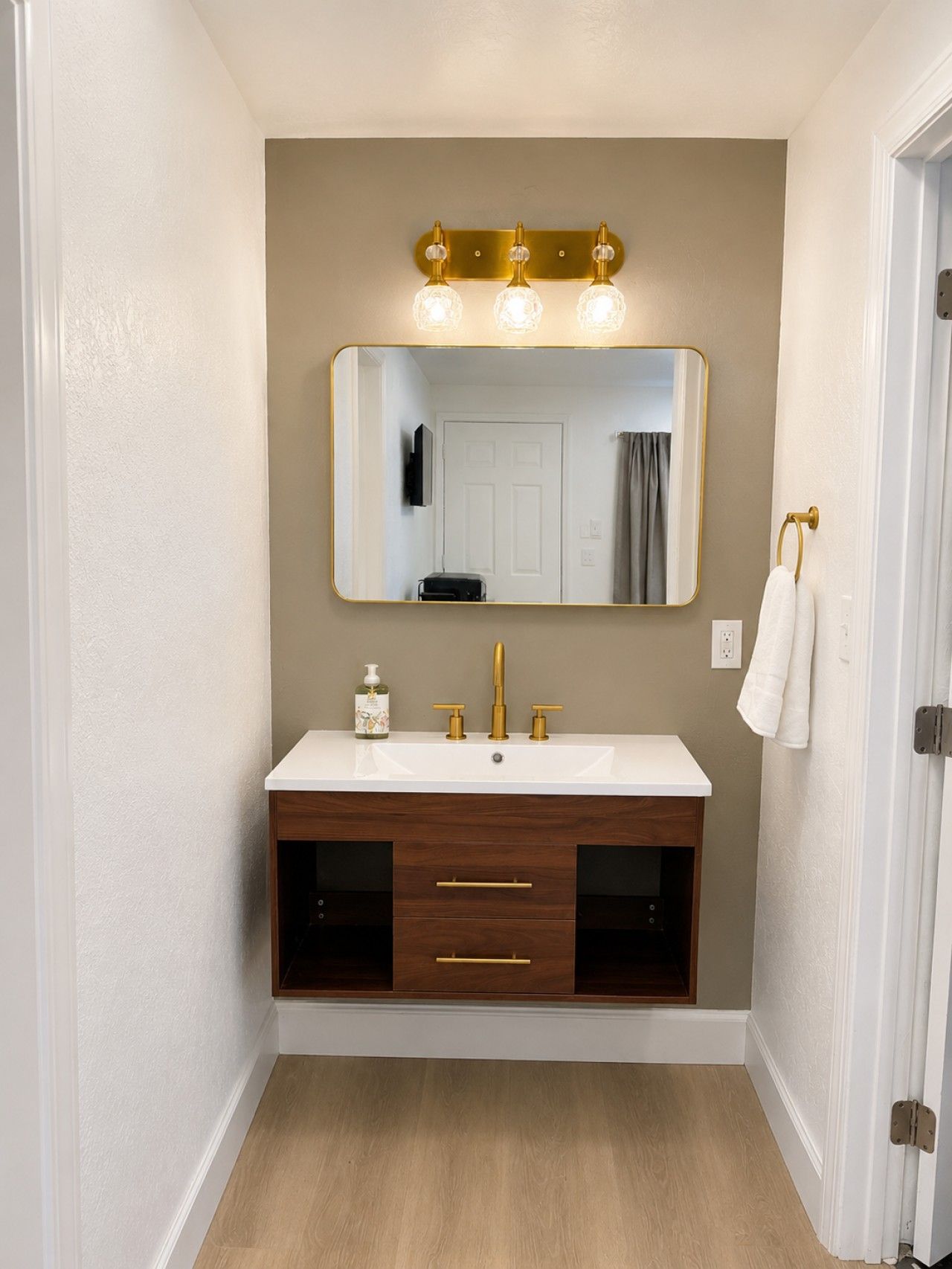 A bathroom with a white vanity, gold fixtures, a mirror with lights, and a towel rack.