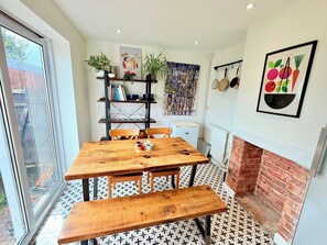 A dining area with a wooden table and benches, a brick fireplace, and a view through a sliding glass door. - Artistic house in Peaceful Kingswood (South Gloucestershire)