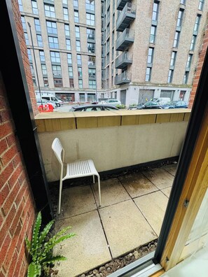 Un balcon avec une chaise en métal blanche, un sol en béton et une vue sur un immeuble en briques doté de nombreuses fenêtres et de balcons.