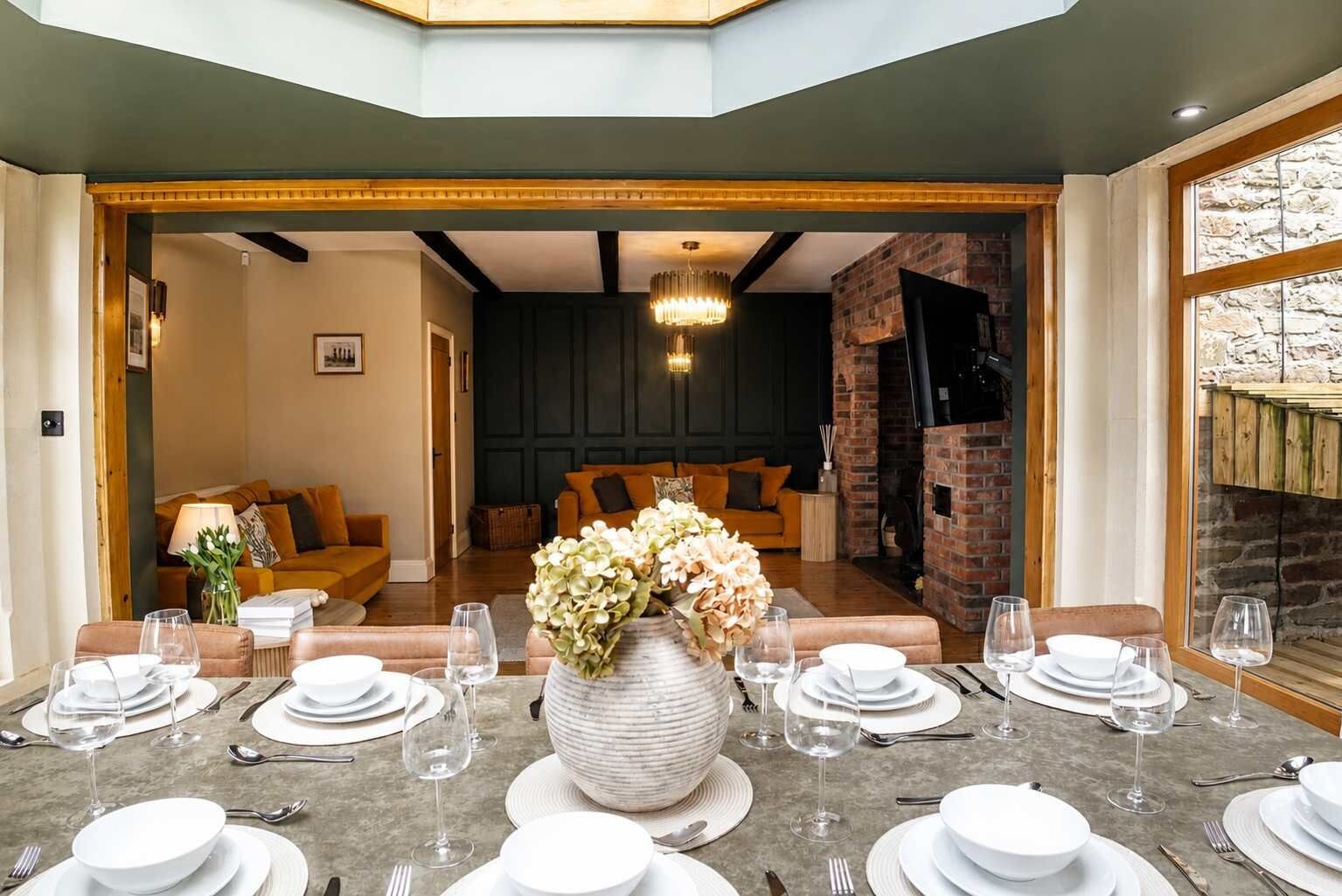 A dining area with a table set for six, a large vase of flowers, and a brick fireplace in the background.