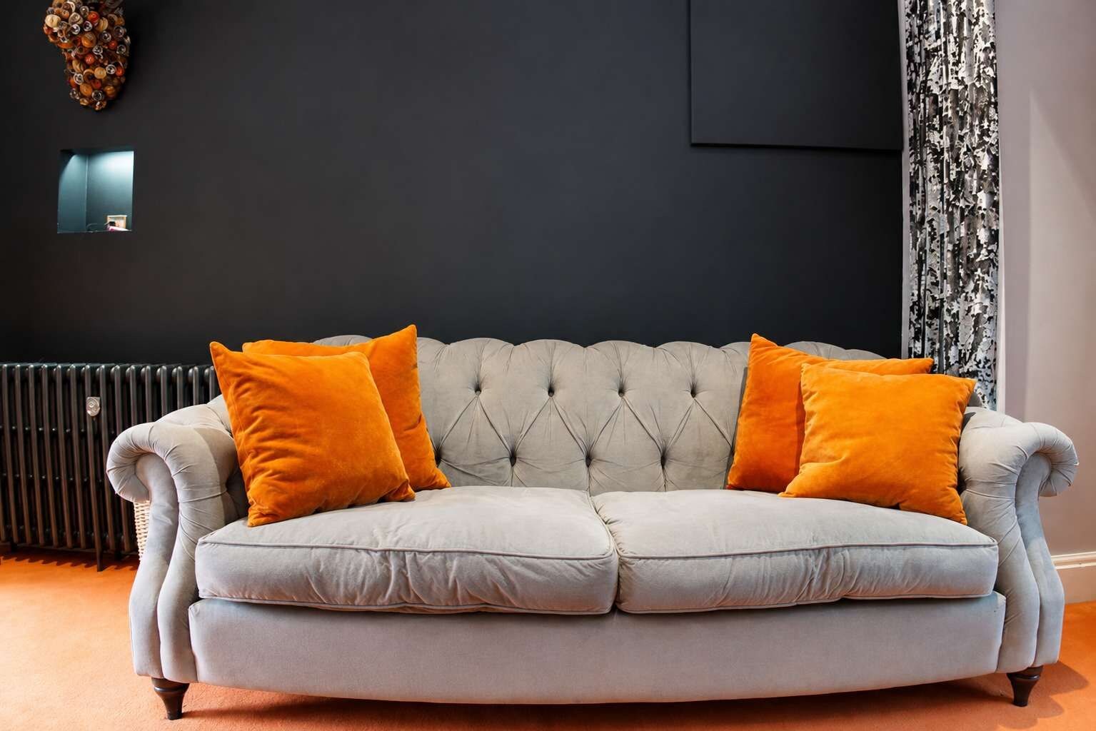 A light grey tufted sofa with two orange cushions, a radiator, a wall-mounted light, and a patterned curtain.